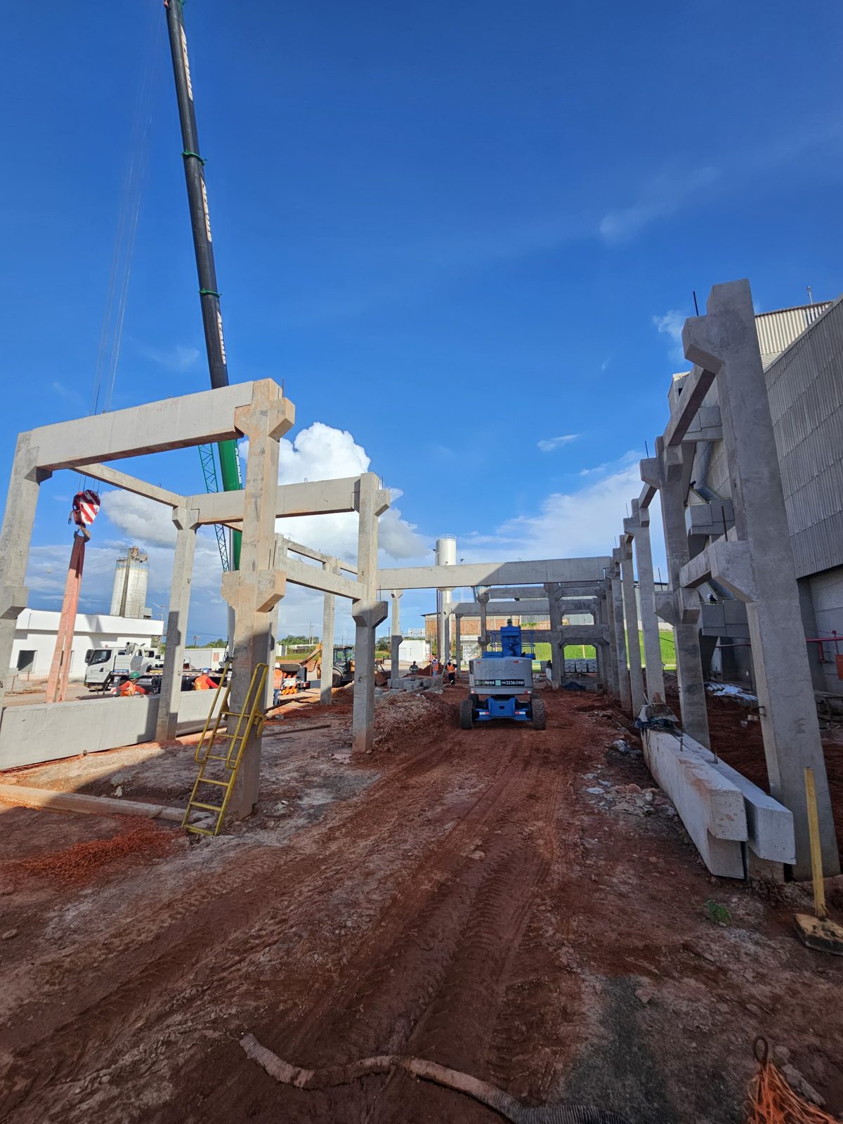 Obra de porte industrial com pórticos pré-fabricados de concreto sendo içados por guindaste